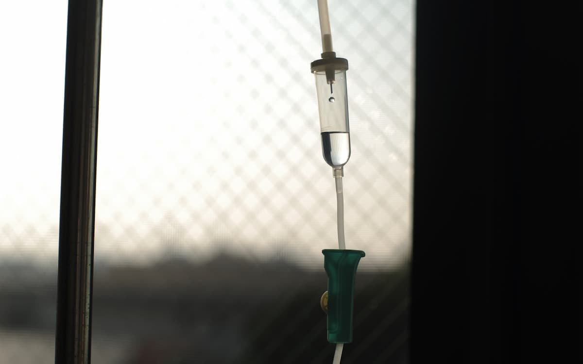 An IV drip chamber and flow clamp backlit by a clinic window